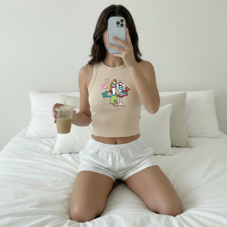 May include: A beige sleeveless ribbed tank top featuring a graphic print of two cartoon characters, Forky and Gabby Gabby, with two pink hearts above them. The top is paired with white shorts that have a subtle, light-colored polka dot pattern.