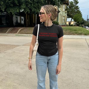 以下が含まれることがあります： 赤い文字で「I SUPPORT WOMEN'S RIGHTS AND WRONGS」と書かれた黒いTシャツ。薄い青色のジーンズ、サングラス、白いハンドバッグを着用。 金色のイヤリングとブレスレットが特徴です。