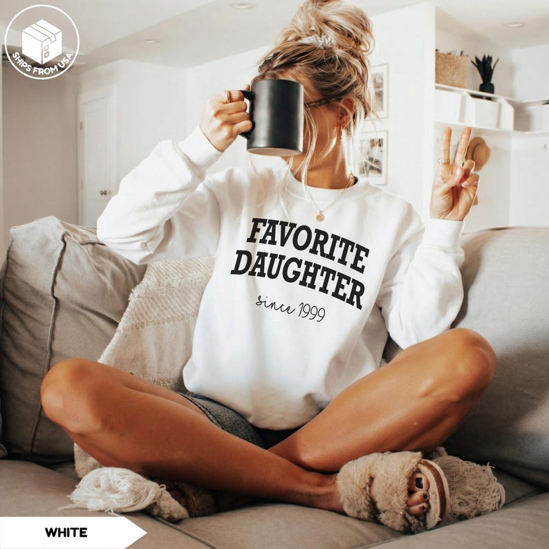 May include: White sweatshirt with the text "FAVORITE DAUGHTER since 1999" in black. The sweatshirt is long-sleeved and has a crew neck. A black mug is held up to the face. The person is sitting on a gray couch.