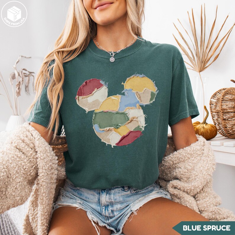 May include: A teal-colored t-shirt featuring a patchwork Mickey Mouse design. The graphic is made of various fabric pieces in red, yellow, blue, and green. The shirt has short sleeves and a relaxed fit. The text "BLUE SPRUCE" is at the bottom right.