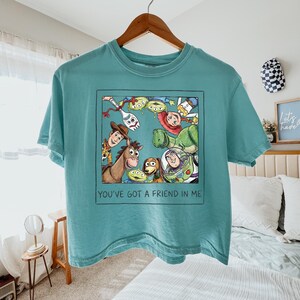May include: A teal t-shirt featuring a square graphic of Toy Story characters including Woody, Buzz Lightyear, Jessie, Rex, Bullseye, Forky, and Aliens. Below the characters, the text reads, "YOU'VE GOT A FRIEND IN ME" in a simple black font.