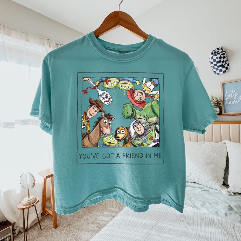 May include: A teal t-shirt featuring a square graphic of Toy Story characters including Woody, Buzz Lightyear, Jessie, Rex, Bullseye, Forky, and Aliens. Below the characters, the text reads, "YOU'VE GOT A FRIEND IN ME" in a simple black font.