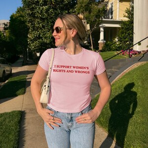 以下が含まれることがあります： 「I SUPPORT WOMEN'S RIGHTS AND WRONGS」の赤い文字が入った、薄いピンク色のTシャツ。半袖で、青いジーンズと合わせています。白いハンドバッグを肩にかけています。