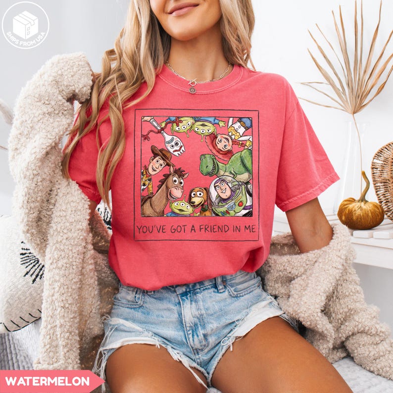 May include: Watermelon-colored t-shirt featuring a framed graphic of animated characters, including a horse, dinosaur, and space ranger. The text "YOU'VE GOT A FRIEND IN ME" is below the graphic. The shirt is paired with denim shorts.