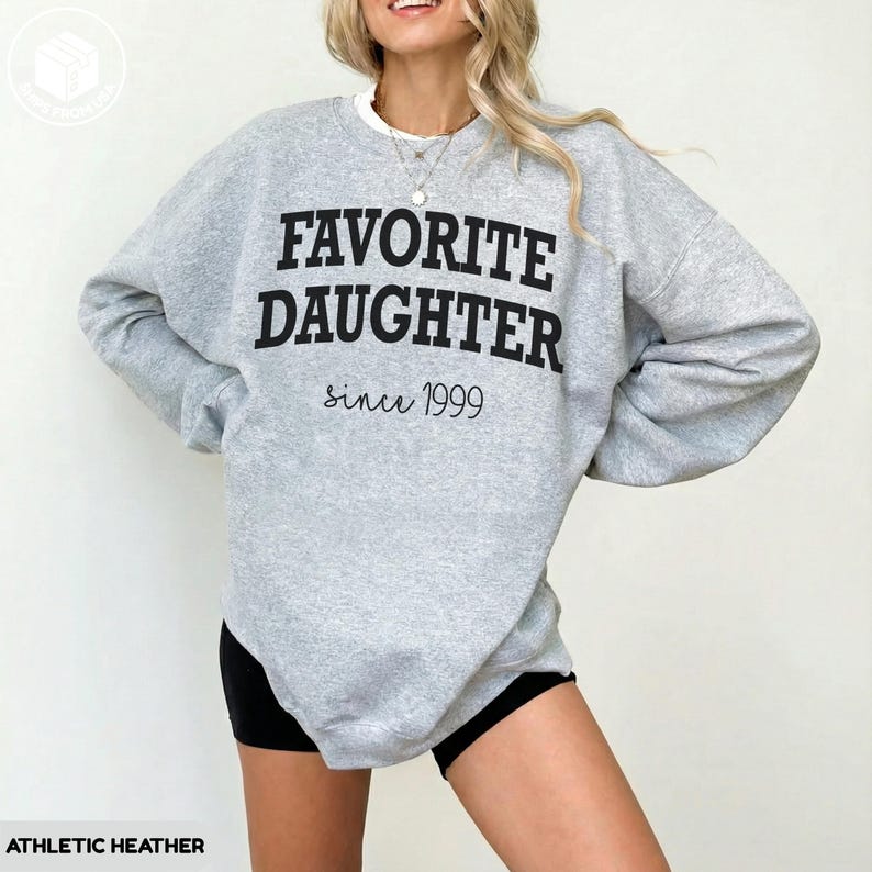 May include: Athletic heather gray sweatshirt with the text "FAVORITE DAUGHTER since 1999" in black. The sweatshirt is a crew neck style and is made of a soft material.
