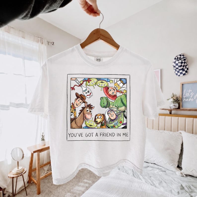 May include: A white short-sleeve t-shirt featuring a square graphic print of Toy Story characters including Woody, Buzz Lightyear, and Forky arranged in a circle. Below the illustration, the text reads, YOU'VE GOT A FRIEND IN ME.
