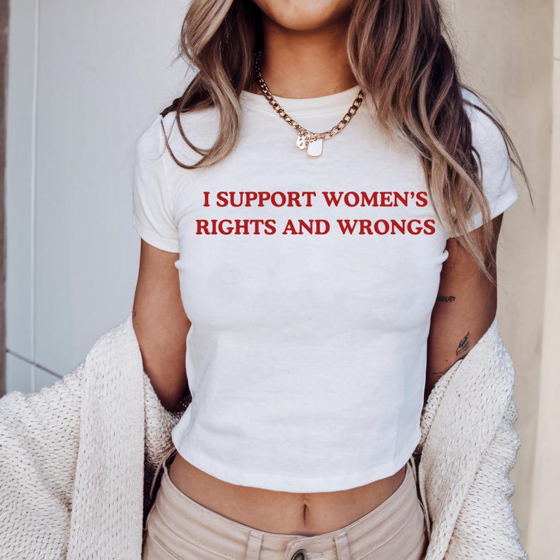以下が含まれることがあります： 赤い文字で「I SUPPORT WOMEN'S RIGHTS AND WRONGS」と書かれた白いクロップトップ。半袖で丸いネックラインです。ゴールドのネックレスが見えます。クリーム色のカーディガンを着ています。