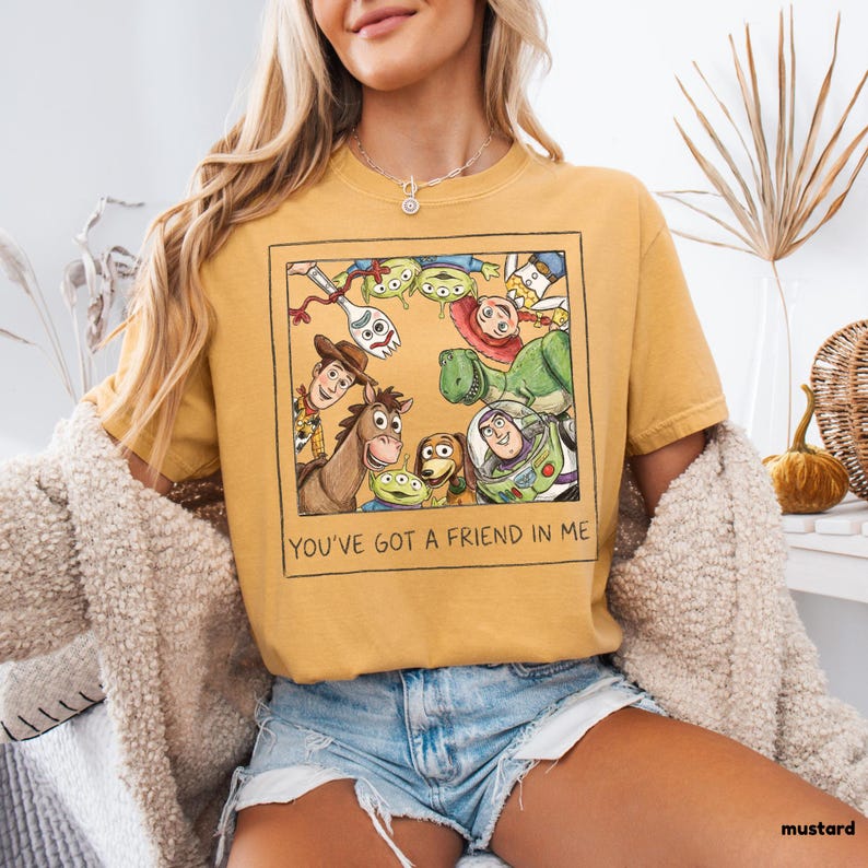 May include: Mustard-colored t-shirt featuring a framed graphic of Toy Story characters, including Woody, Buzz Lightyear, and the Little Green Men. The text "YOU'VE GOT A FRIEND IN ME" is printed below the graphic. The shirt is a casual style.