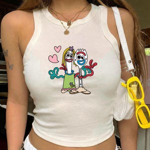 May include: A white sleeveless tank top featuring a graphic print of two anthropomorphic utensil characters with googly eyes, one wearing a green dress and the other a red headband, surrounded by pink hearts and red squiggly lines.