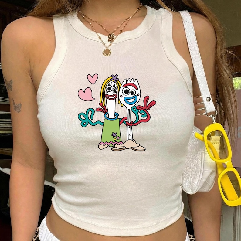 May include: A white sleeveless tank top featuring a graphic print of two anthropomorphic utensil characters with googly eyes, one wearing a green dress and the other a red headband, surrounded by pink hearts and red squiggly lines.
