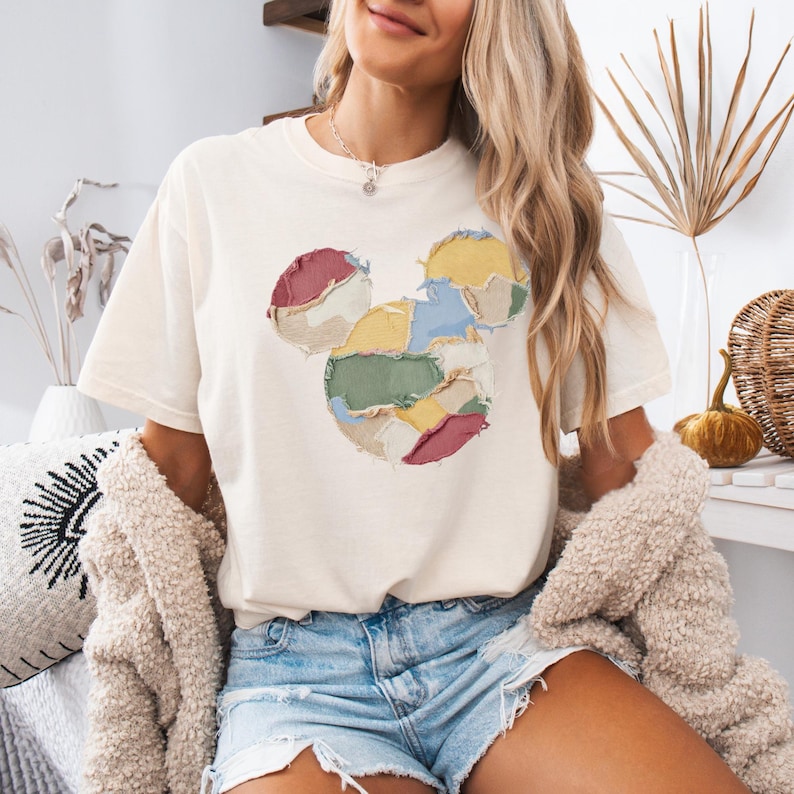 May include: Cream-colored t-shirt featuring a Mickey Mouse design made of colorful fabric patches. The design includes red, yellow, green, and blue patches. The shirt is paired with denim shorts and a beige cardigan.