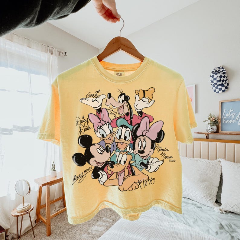 May include: Yellow t-shirt featuring a graphic of Disney characters including Mickey Mouse, Minnie Mouse, Donald Duck, Daisy Duck, Goofy, and Pluto. The shirt has signatures and the words "Mickey Mouse" and "Minnie Mouse" on it.