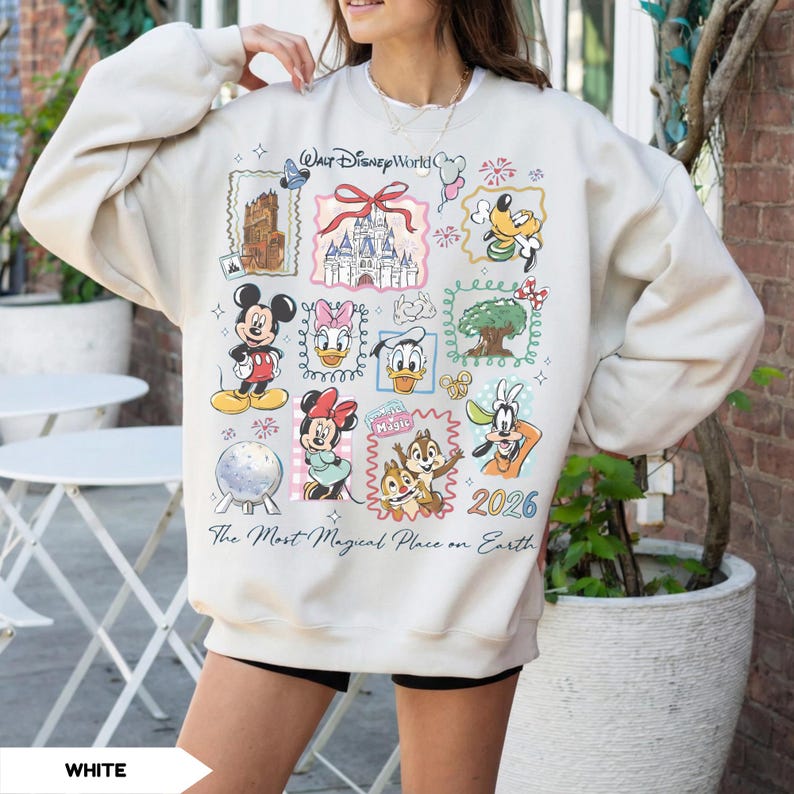 Puede incluir: Sudadera de color crema con un colorido collage de personajes y lugares emblem&aacute;ticos de Disney. El dise&ntilde;o incluye a Mickey Mouse, Minnie Mouse, Donald Duck y Goofy, junto con el texto "Walt Disney World" y "The Most Magical Place on Earth". Tambi&eacute;n se muestra el a&ntilde;o "2026".