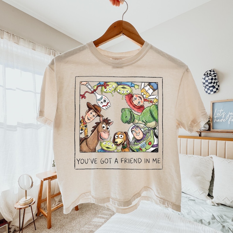 May include: A cream-colored short-sleeve t-shirt featuring a square graphic print of Toy Story characters including Woody, Buzz Lightyear, Rex, and Forky. The illustration is framed like a Polaroid photo with the text YOU'VE GOT A FRIEND IN ME.
