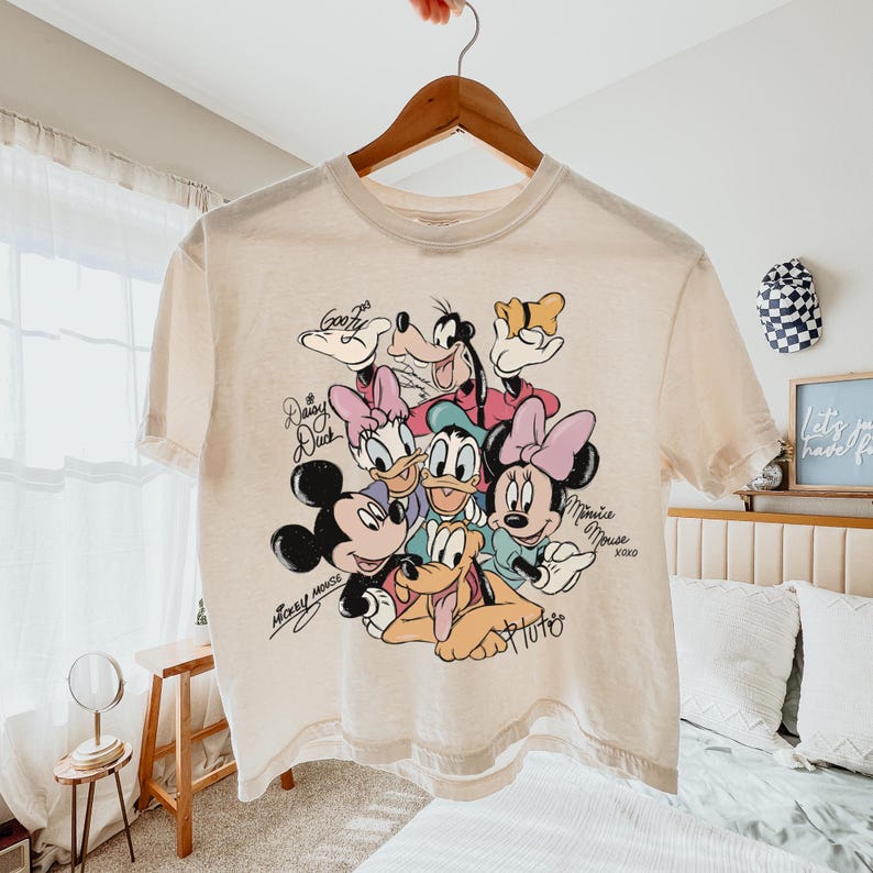 May include: Cream-colored t-shirt featuring a colorful graphic of Disney characters, including Mickey Mouse, Minnie Mouse, Donald Duck, Daisy Duck, Goofy, and Pluto. The shirt has signatures and the words "Mickey Mouse" and "Minnie Mouse" printed on it.