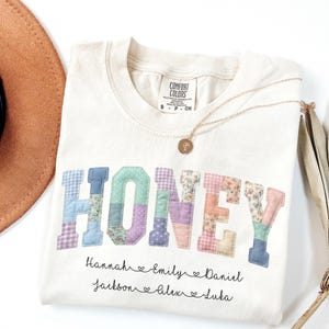 May include: Cream-colored t-shirt with the word "HONEY" in patchwork letters. The letters are in various pastel colors and patterns. Below the word are names. A gold necklace and a brown hat are in the background.