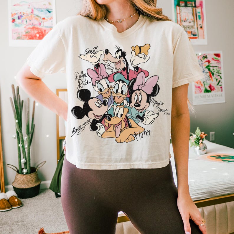 May include: Cream-colored t-shirt featuring a graphic of Disney characters including Mickey Mouse, Minnie Mouse, Donald Duck, Daisy Duck, Goofy, and Pluto. The shirt has short sleeves and a crew neck. The characters' names are written in cursive.