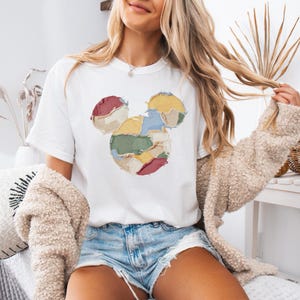 May include: White t-shirt featuring a Mickey Mouse design. The design is made of colorful, patchwork-style fabric pieces in shades of red, yellow, green, and blue. The shirt has a classic crew neck and short sleeves.