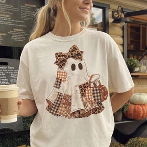 May include: A cream-colored short-sleeve t-shirt featuring a central graphic of a ghost adorned with a leopard print bow. The ghost is composed of various fabric patches in brown, orange, and white plaid, floral, and leopard patterns, holding a pumpkin.
