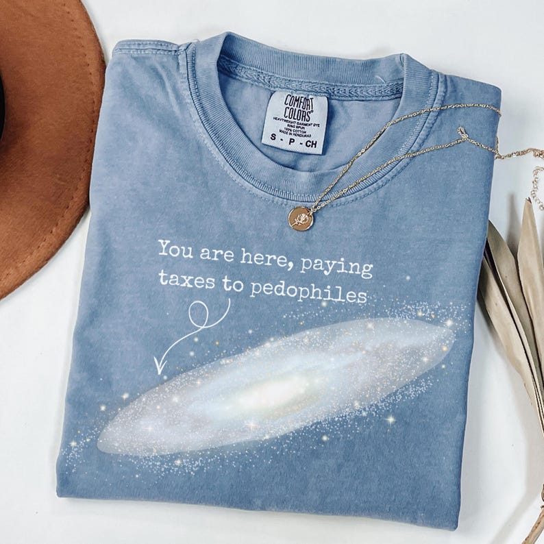 Puede incluir: Una camiseta azul Comfort Colors con el texto "You are here, paying taxes to pedophiles" y un gr&aacute;fico de galaxia. Un collar dorado con un colgante est&aacute; sobre la camiseta.