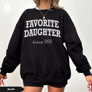 May include: Black sweatshirt with the text "FAVORITE DAUGHTER" in bold white letters, with "since 1999" in a cursive font. The sweatshirt has a white collar and is a casual style.