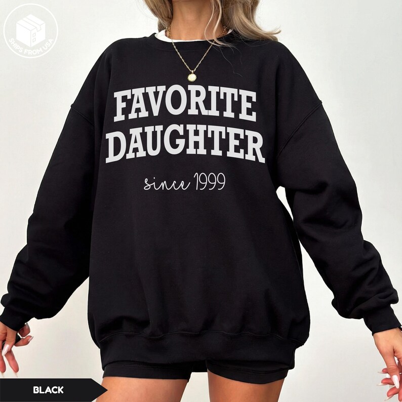 May include: Black sweatshirt with the text "FAVORITE DAUGHTER" in bold white letters, with "since 1999" in a cursive font. The sweatshirt has a white collar and is a casual style.