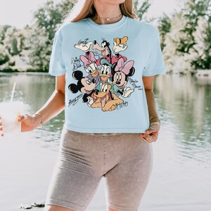 May include: A light blue t-shirt featuring a graphic of Mickey Mouse and friends, including Minnie Mouse, Donald Duck, and Goofy. The characters are in a colorful design with their names written above them. The shirt is a crew neck style.