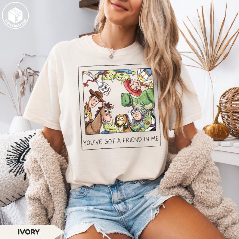 May include: Ivory-colored t-shirt featuring a framed graphic of characters from the animated film Toy Story. The graphic includes Woody, Buzz Lightyear, Rex, and the Little Green Men. The text "YOU'VE GOT A FRIEND IN ME" is printed below the graphic.