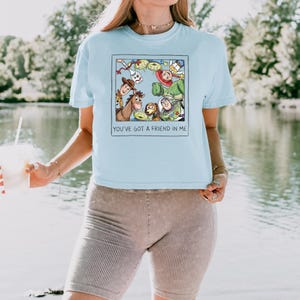 May include: A light blue t-shirt featuring a square graphic print of various animated characters from Toy Story arranged in a circle. Below the characters, the text reads, "YOU'VE GOT A FRIEND IN ME." The shirt has short sleeves and a crew neckline.