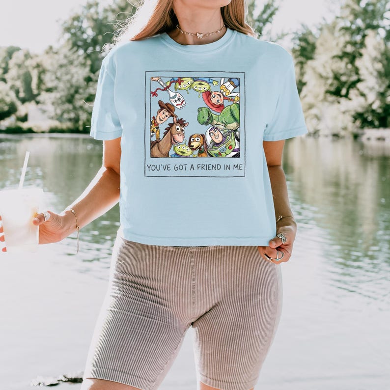 May include: A light blue t-shirt featuring a square graphic print of various animated characters from Toy Story arranged in a circle. Below the characters, the text reads, "YOU'VE GOT A FRIEND IN ME." The shirt has short sleeves and a crew neckline.