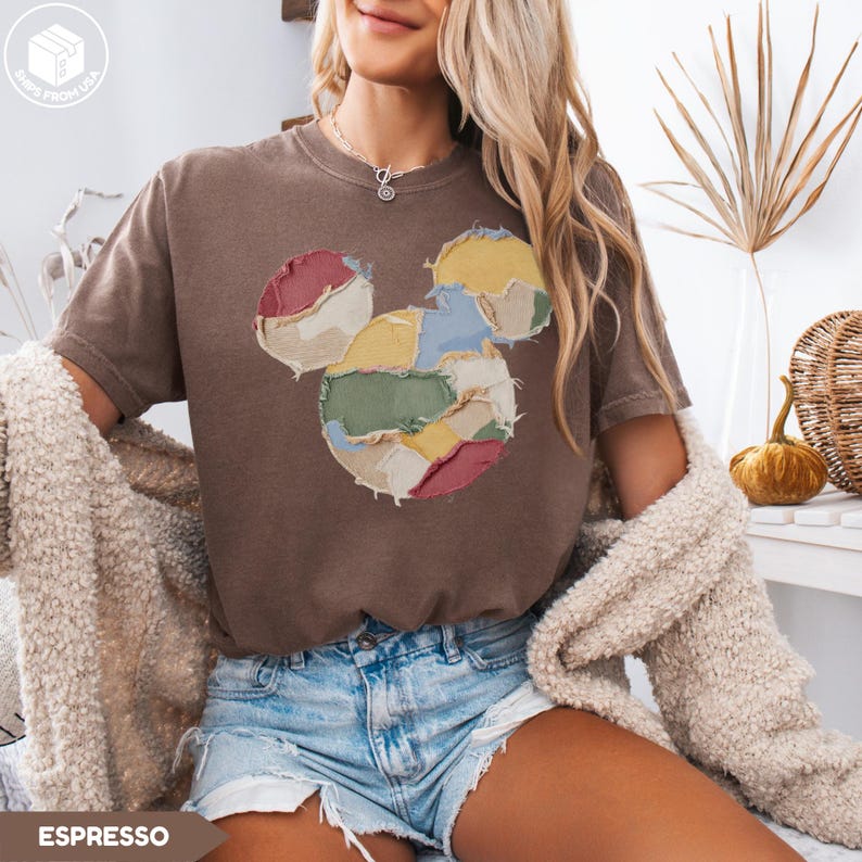 May include: Espresso-colored t-shirt featuring a patchwork Mickey Mouse design in various colors. The shirt has a relaxed fit and short sleeves. The image also shows denim shorts and a cream-colored cardigan.