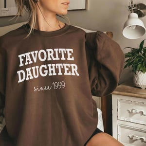 May include: A brown sweatshirt with the words "FAVORITE DAUGHTER" in bold white letters, and "since 1999" in a cursive font. The sweatshirt has a crew neck and long sleeves.