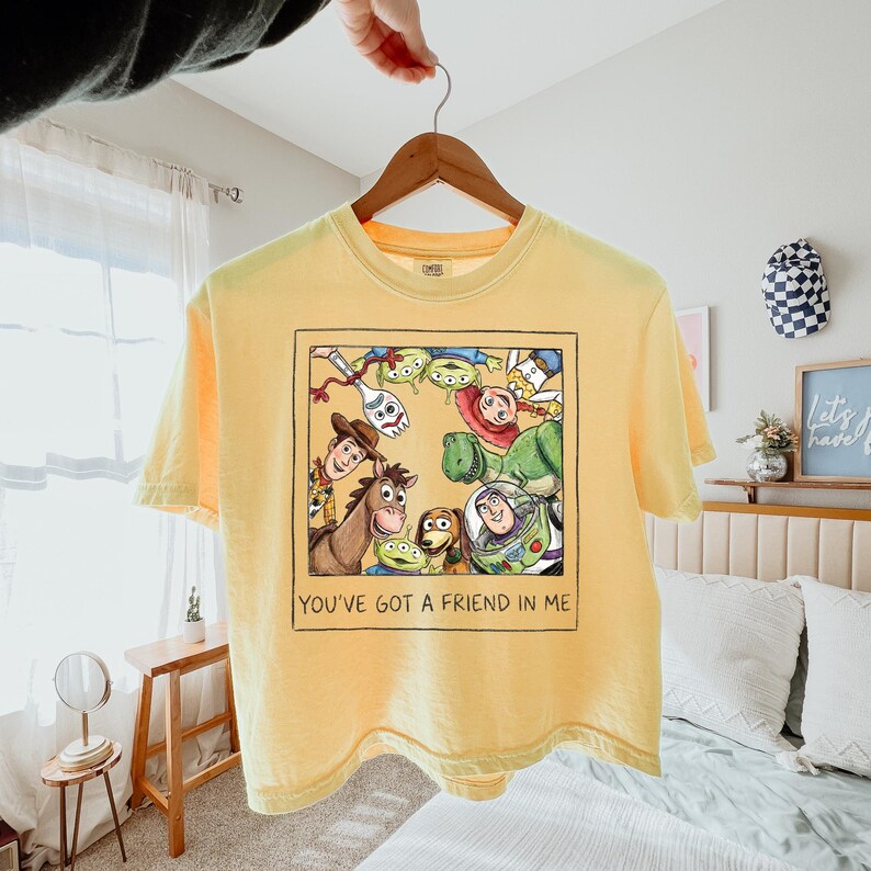 May include: A pale yellow short-sleeve t-shirt featuring a square graphic print of various animated toy characters arranged in a circle. Below the illustration, the text reads, "YOU'VE GOT A FRIEND IN ME" in a simple, dark, hand-drawn style font.