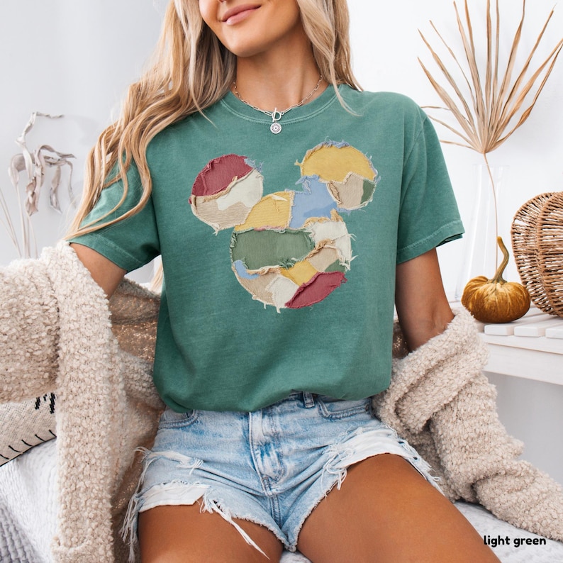 May include: Light green t-shirt featuring a patchwork Mickey Mouse design. The Mickey Mouse design is made of various colors, including red, yellow, blue, and green. The shirt is a comfortable, casual style.