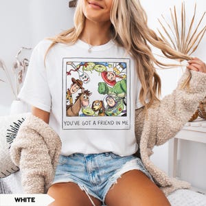 May include: White t-shirt featuring a framed illustration of characters from the animated film Toy Story. The illustration includes Woody, Buzz Lightyear, and other characters. The text "YOU'VE GOT A FRIEND IN ME" is below the illustration.