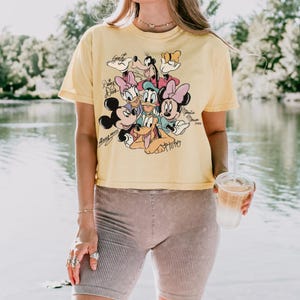 May include: Yellow short-sleeved t-shirt featuring a graphic of Disney characters including Mickey Mouse, Minnie Mouse, Donald Duck, and Goofy. The shirt has a vintage look and is paired with ribbed shorts. The image is taken outdoors near a body of water.