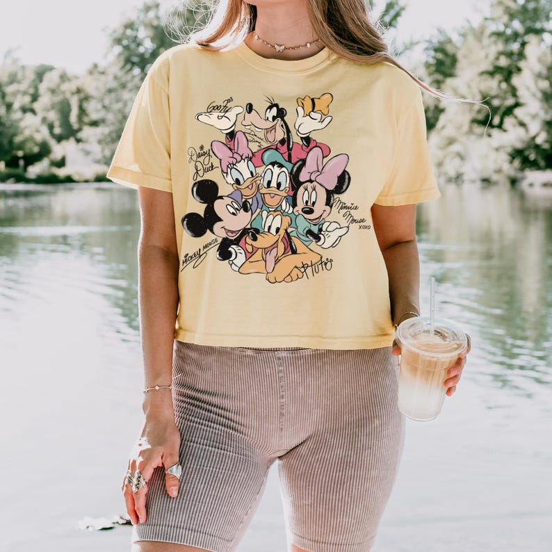 May include: Yellow short-sleeved t-shirt featuring a graphic of Disney characters including Mickey Mouse, Minnie Mouse, Donald Duck, and Goofy. The shirt has a vintage look and is paired with ribbed shorts. The image is taken outdoors near a body of water.