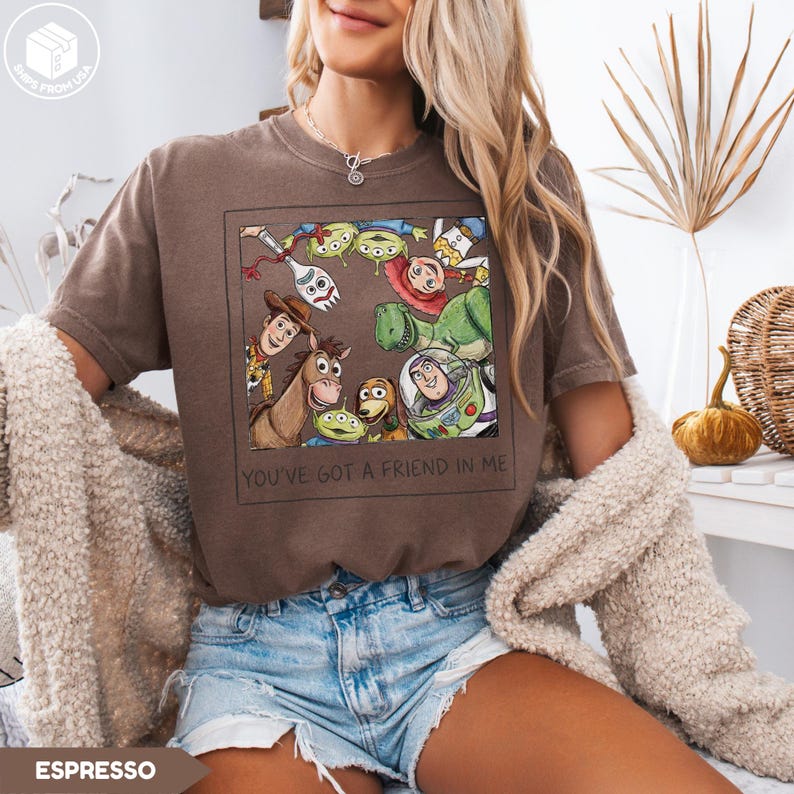 May include: Espresso-colored t-shirt featuring a framed graphic of Toy Story characters, including Woody, Buzz Lightyear, and the aliens. The text "YOU'VE GOT A FRIEND IN ME" is printed below the image. The shirt is worn by a person.