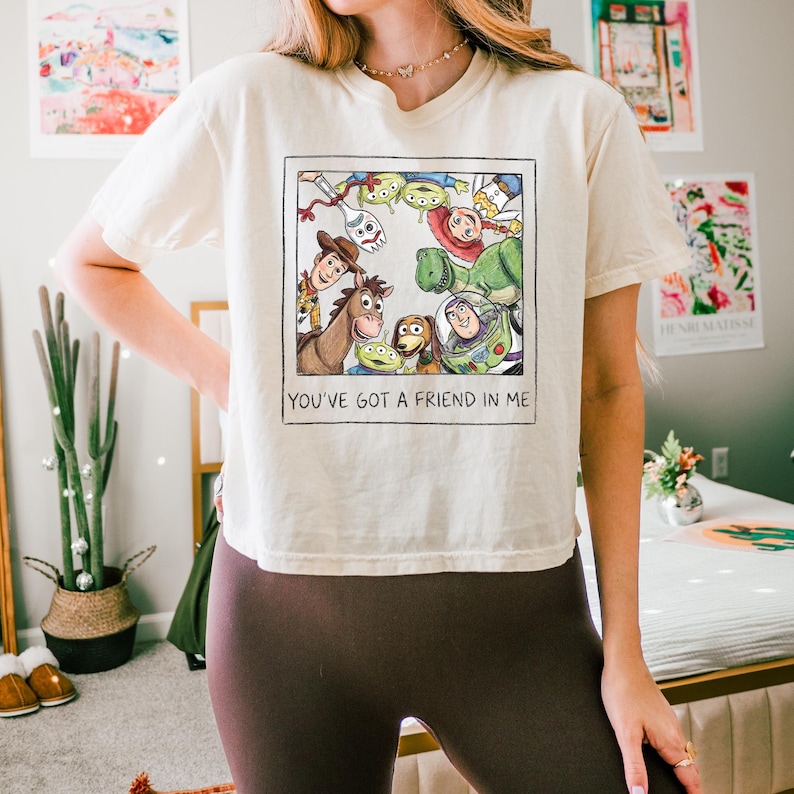 May include: A cream-colored short-sleeve t-shirt featuring a square graphic print of Toy Story characters including Woody, Buzz Lightyear, and Rex. Below the illustration, the text reads YOU'VE GOT A FRIEND IN ME in a simple black font.
