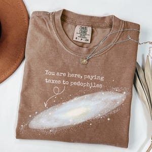 Puede incluir: Camiseta Comfort Colors marr&oacute;n con el texto "You are here, paying taxes to pedophiles" y un gr&aacute;fico de galaxia. Un collar plateado con un colgante est&aacute; sobre la camiseta. La camiseta es talla S-P-CH.