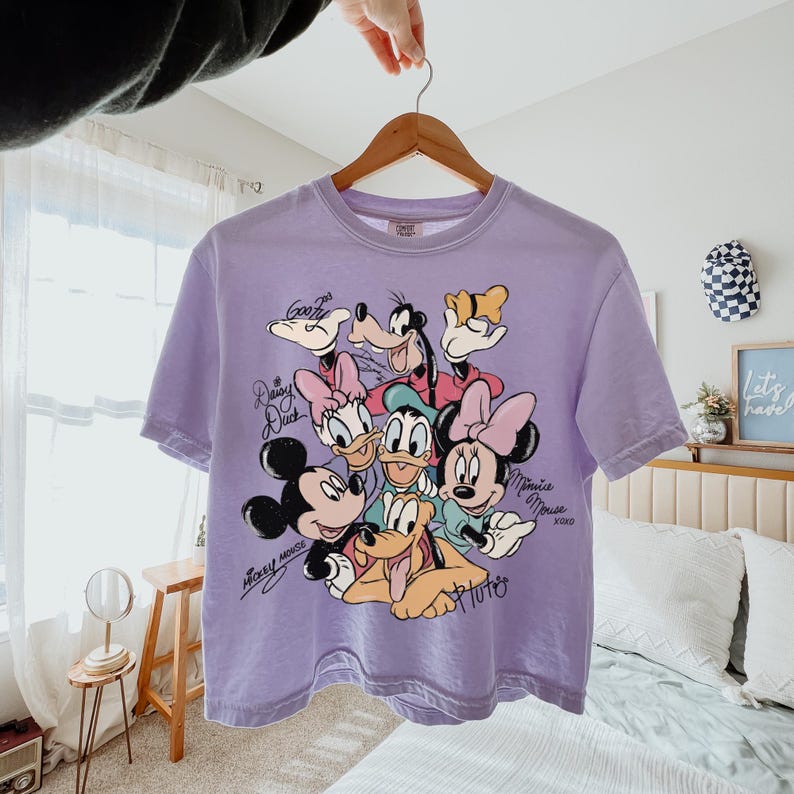 May include: A lilac-colored t-shirt featuring a graphic of Disney characters including Mickey Mouse, Minnie Mouse, Donald Duck, Daisy Duck, Goofy, and Pluto. The shirt has short sleeves and a crew neck. The characters' names are written in a script font.