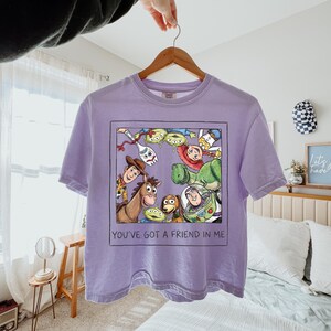 May include: A light purple short-sleeve t-shirt featuring a square graphic print of Toy Story characters including Woody, Buzz Lightyear, Rex, Jessie, and Forky. The text "YOU'VE GOT A FRIEND IN ME" is printed in black below the character illustration.
