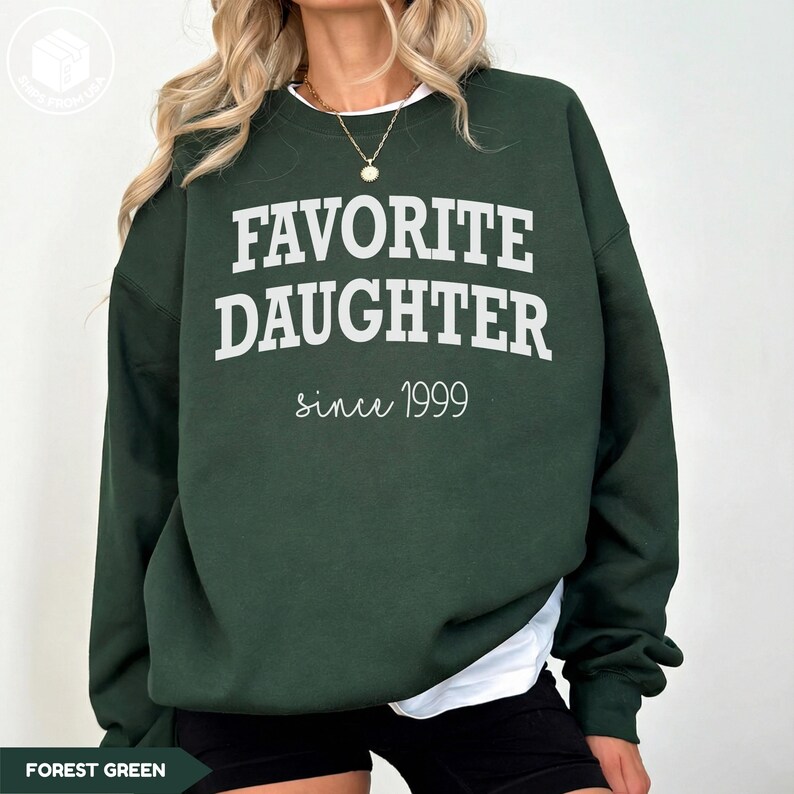 May include: Forest green sweatshirt with the text "FAVORITE DAUGHTER since 1999" in white. The sweatshirt is a solid color and has a crew neck. The text is centered on the front of the sweatshirt.
