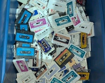 May include: A large collection of assorted audio cassette tapes housed within a blue plastic bin. The tapes feature various colored labels, including white, blue, yellow, and purple, with printed text such as Secrets of the Universe and Real Magic.