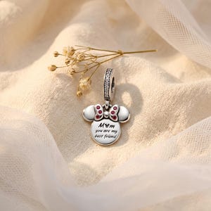 Puede incluir: Un charm plateado con forma de cabeza de rat&oacute;n y un lazo con peque&ntilde;as piedras rojas. El charm tiene una anilla incrustada con piedras transparentes y est&aacute; grabado con el texto "Mom you are my best friend" junto a un icono de cabeza de rat&oacute;n.
