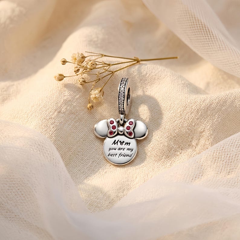 Puede incluir: Un charm plateado con forma de cabeza de rat&oacute;n y un lazo con peque&ntilde;as piedras rojas. El charm tiene una anilla incrustada con piedras transparentes y est&aacute; grabado con el texto "Mom you are my best friend" junto a un icono de cabeza de rat&oacute;n.