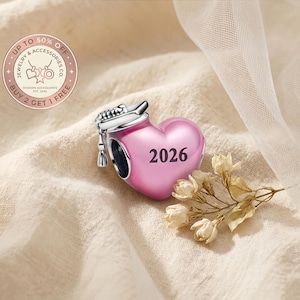 Può includere: Un charm a forma di cuore rosa con l'anno "2026" stampato sopra. Il charm ha una parte superiore color argento con una nappa. Il charm fa parte di una collezione di gioielli e accessori.
