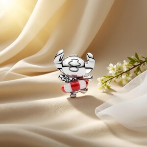 May include: A silver-toned metal charm featuring a stylized character with large ears and a head marking, wearing a red and white striped life preserver ring around its waist. The charm has a polished, reflective surface and a smooth, rounded design.