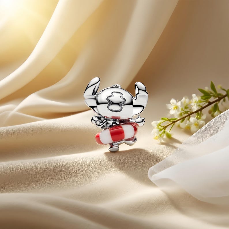 May include: A silver-toned metal charm featuring a stylized character with large ears and a head marking, wearing a red and white striped life preserver ring around its waist. The charm has a polished, reflective surface and a smooth, rounded design.