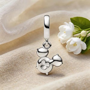 May include: A silver charm pendant in the shape of a cartoon character with a clear crystal center. The charm has a loop for attaching to a bracelet or necklace. White flowers and green leaves are in the background.